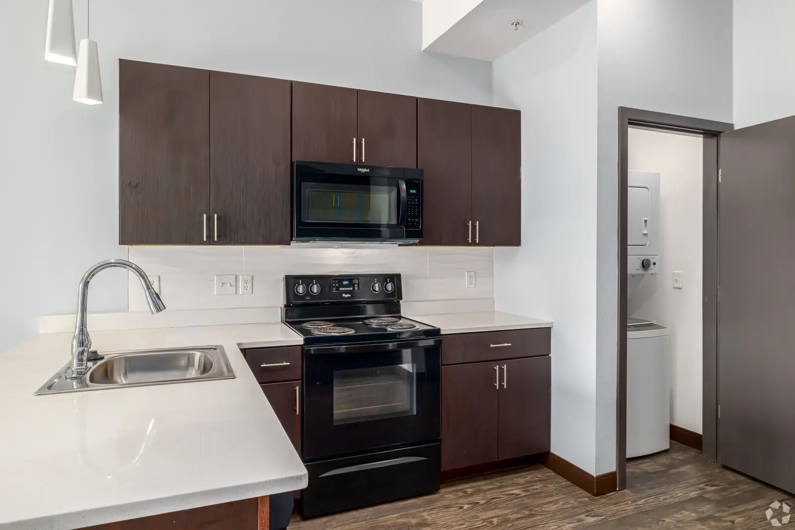 Modern apartment kitchen with dark wood cabinetry, white countertops, black appliances, and an in-unit washer and dryer.